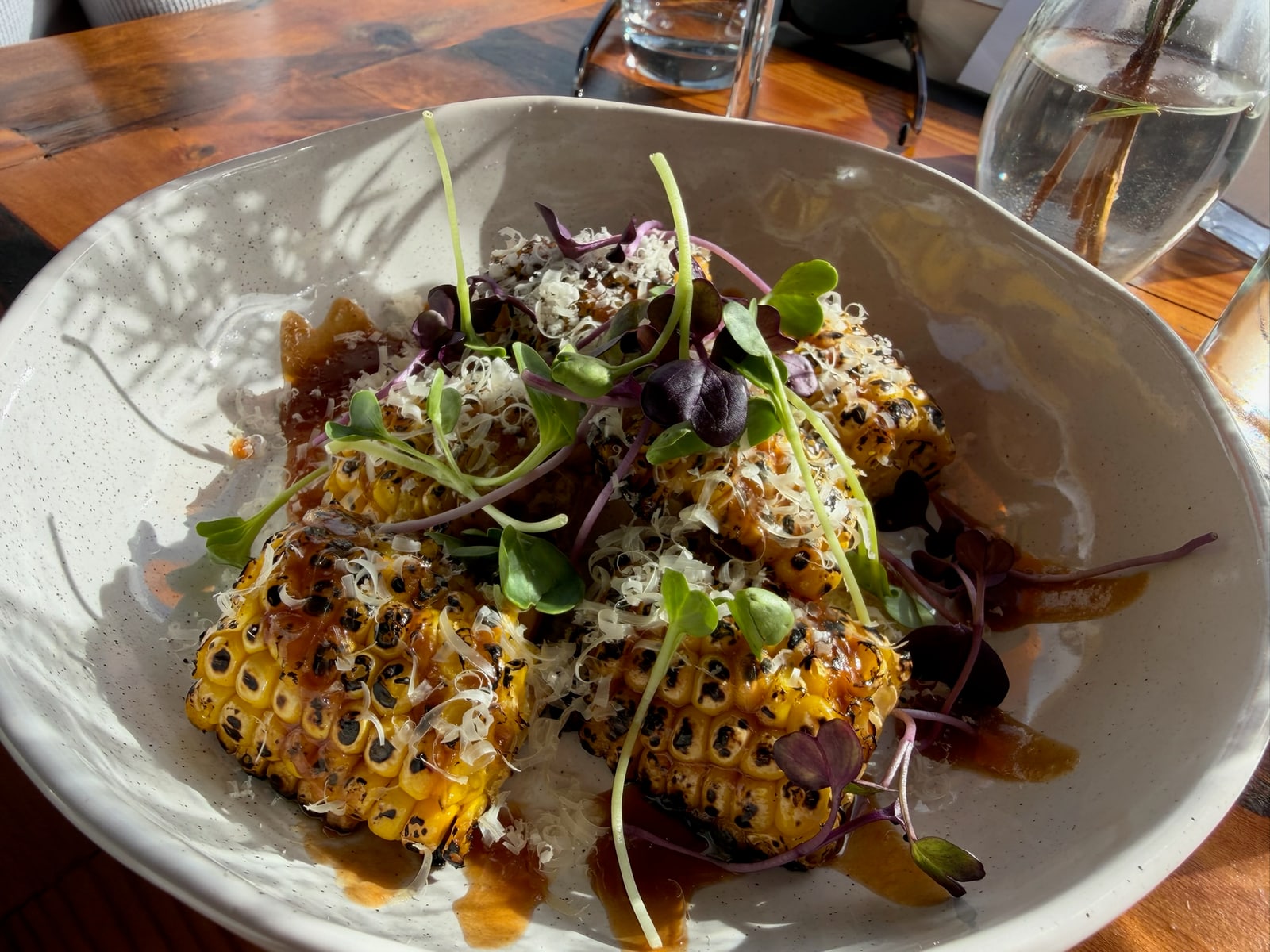 Grilled corn topped with microgreens and shaved cheese in a white bowl on a sunlit wooden table.